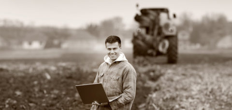 Young landowner with laptop supervising work on farmland, tractor in background, black and white image